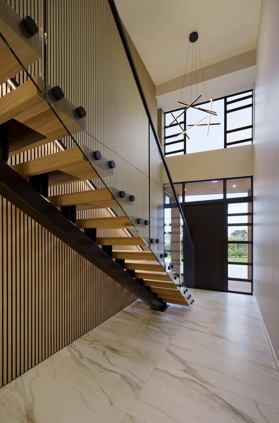 Double-height entranceway with a floating timber stair, glass balustrade and full-height windows either side of the front door