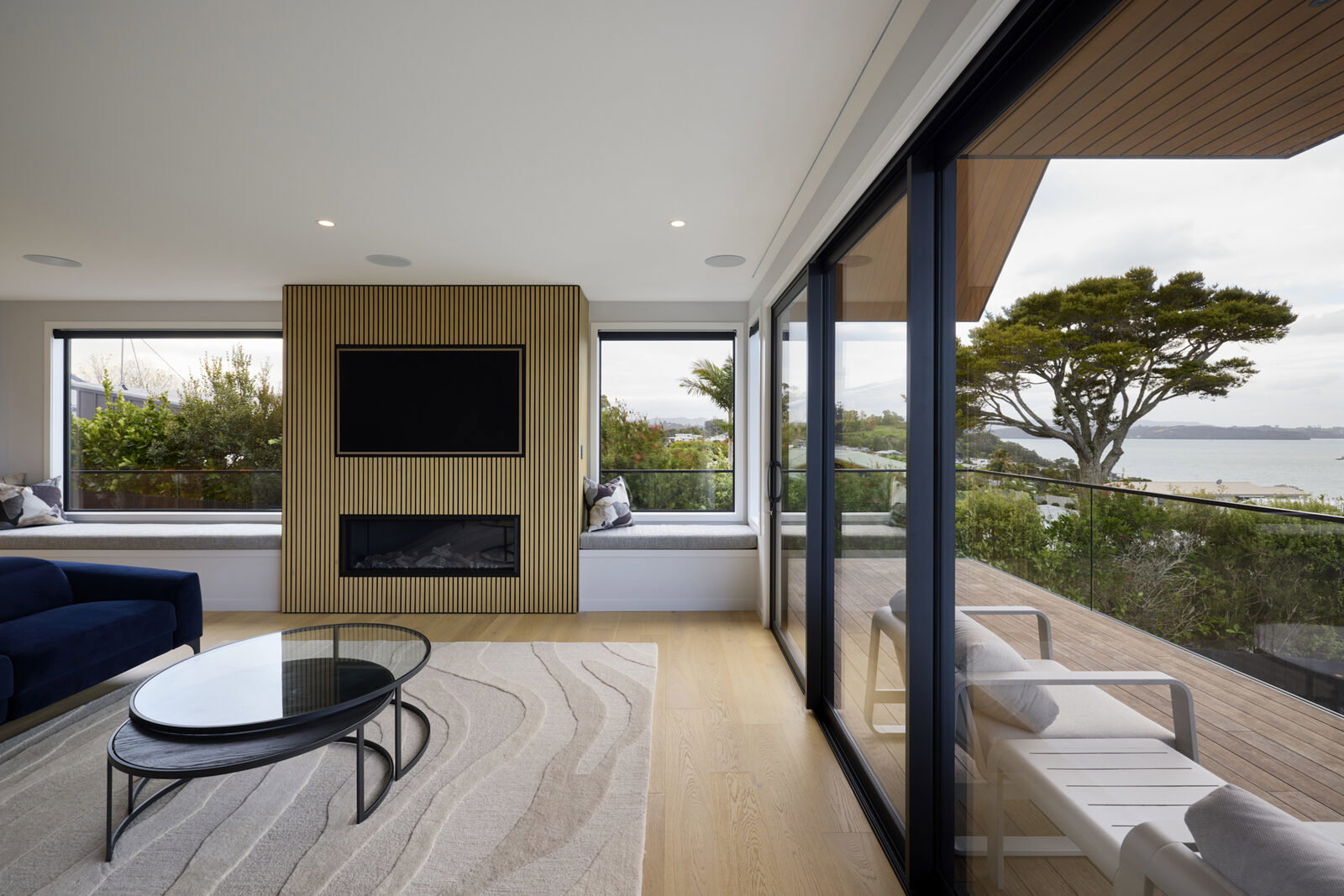 Living room with a slatted timber fireplace feature wall and window seats, sliding glass doors open to the deck and sea view beyond